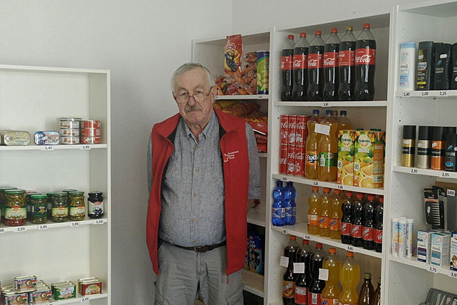 Happy Birthday, Kiosk! Ein DRK-Mitarbeiter zwischen weißen Regalen, die mit Konserven, Getränken und Hygieneartikeln gefüllt sind.