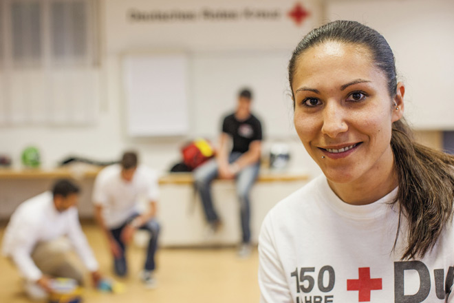 Eine junge Frau mit DRK-T-Shirt im Vordergrund, im Hintergrund eine kleine Gruppe von Menschen