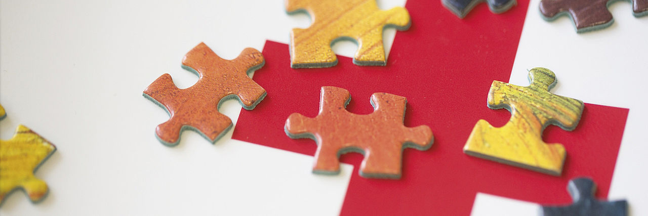 Rotes Kreuz auf weißem Grund, darauf verstreute Puzzle-Teile