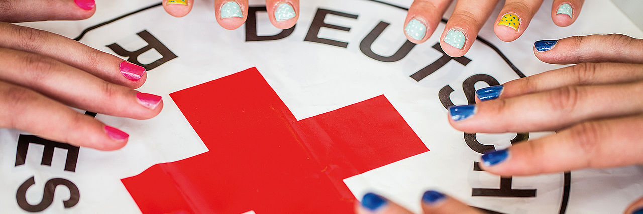 Viele Hände mit bunt lackierten Fingernägeln gruppieren sich um das DRK-Logo